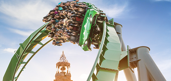 Ao centro da imagem, vê-se pessoas com os braços levantados dentro de um carrinho em movimento em uma montanha-russa, The Incredible Hulk Coaster®.