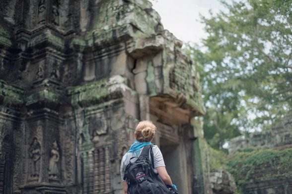 Uma mulher viajante está de costas com um coque e uma mochila preta enquanto observa um templo antigo em ruínas com uma árvore ao fundo