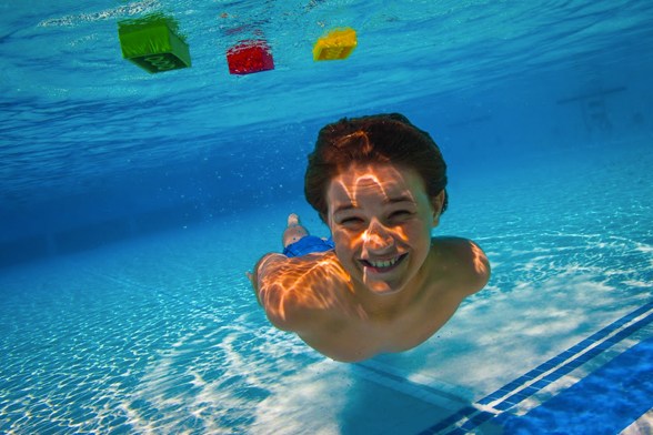 Um menino sorri embaixo d'água, em uma piscina com blocos LEGO® boiando acima dele.