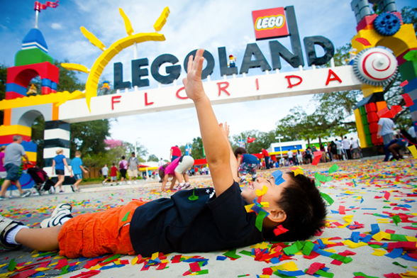 Um menino sorri e se diverte com papeis coloridos que estão espalhados por todo o chão. Ao fundo, se vê a entrada principal do LEGOLAND® Florida Resort, em Orlando.