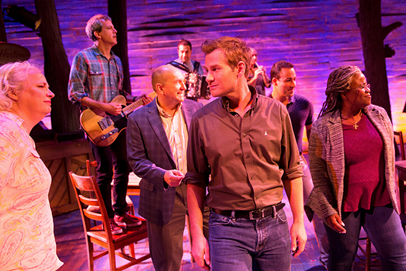 Cena do musical Come From Away, da Broadway. O palco está iluminado em uma tonalidade de rosa, os músicos estão ao fundo, em pé em cima de cadeiras, e os atores à frente.