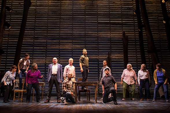 Cena do musical Come From Away, da Broadway, 23 atores olham para o horizonte, em um cenário simples composto de uma mesa e cadeiras.