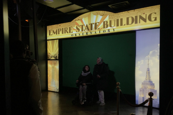 Duas pessoas posam para foto em frente a um fundo verde, com uma borda que diz "Empire State Building" (Foto: Natália Grandi)