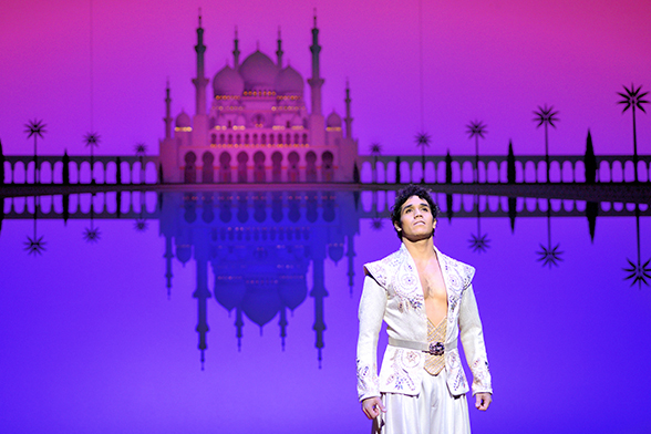 Aladdin no musical homônimo da Broadway, em Nova York. Ao fundo, um castelo e cores rosa e azuis.